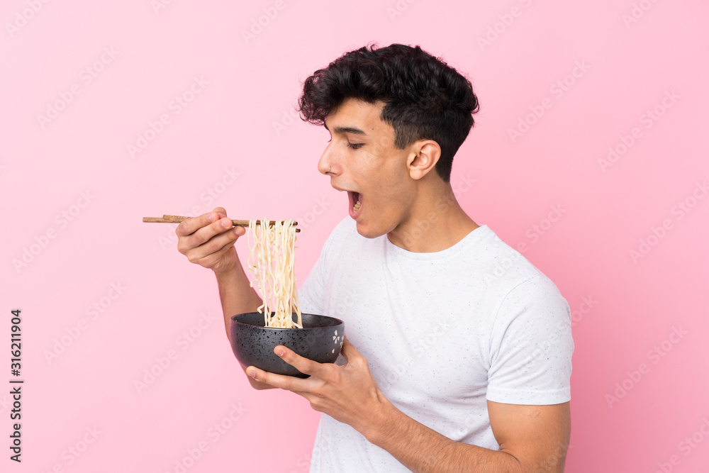 Young Argentinian man over isolated white background holding a bowl of noodles with chopsticks and eating it Young Argentinian man over isolated white background holding a bowl of noodles with chopsticks and eating it