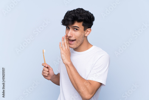Young Argentinian man brushing his teeth over isolated blue background whispering something