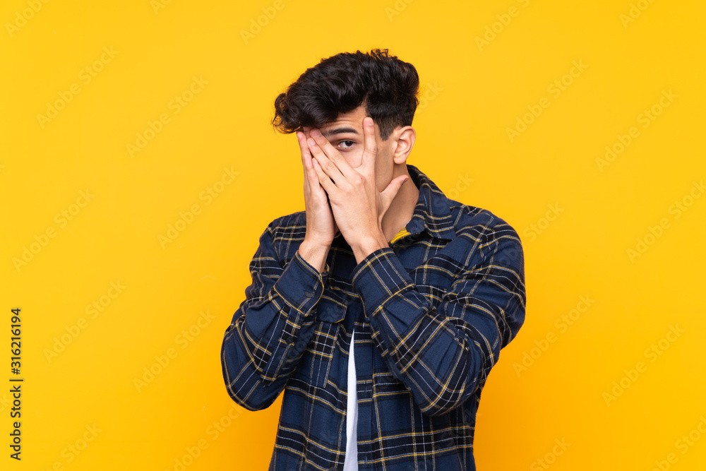 Young Argentinian man over isolated yellow background covering eyes and looking through fingers Young Argentinian man over isolated yellow background covering eyes and looking through fingers
