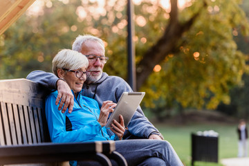Smiling senior active couple sitting on the bench looking at tablet computer. Using modern technology by elderly.
