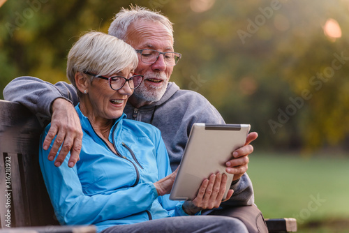 Smiling senior active couple sitting on the bench looking at tablet computer. Using modern technology by elderly.