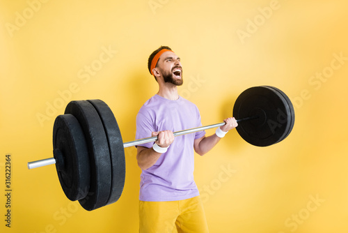 emotional bearded sportsman working out with barbell on yellow