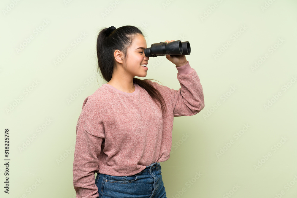 Young teenager Asian girl over isolated green background with black binoculars Young teenager Asian girl over isolated green background with black binoculars