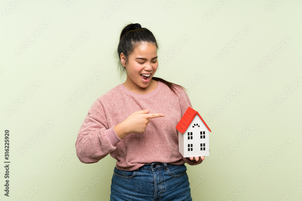 Young teenager Asian girl over isolated green background holding a little house Young teenager Asian girl over isolated green background holding a little house