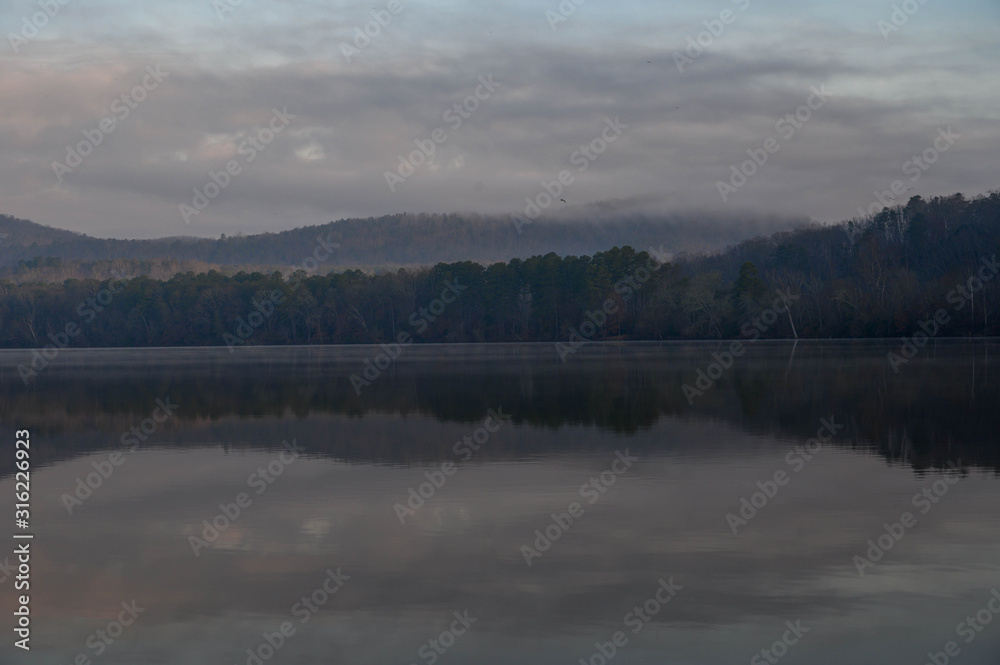 Fototapeta premium The reflections of the rolling mountains on a cloudy early morning. The haze over the water on a cold winter sunrise morning.