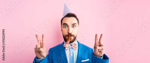 panoramic shot of businessman in party cap holding party blower in mouth and showing peace sign isolated on pink photo