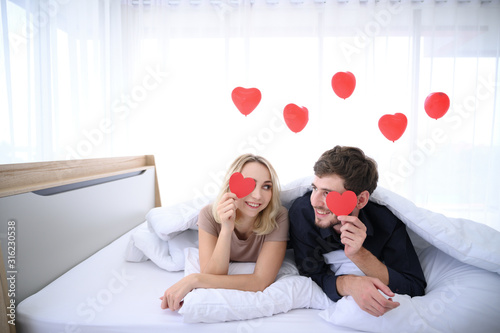 Couple. Love. Valentine's day. Man and woman are holding red paper hearts and look at each other with smiling on bed