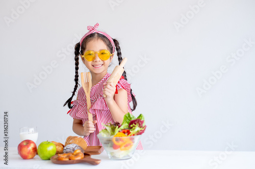 Cute Asian girl shows kitchenware with various types of fruit and vegetables and stand in front of white wall background.