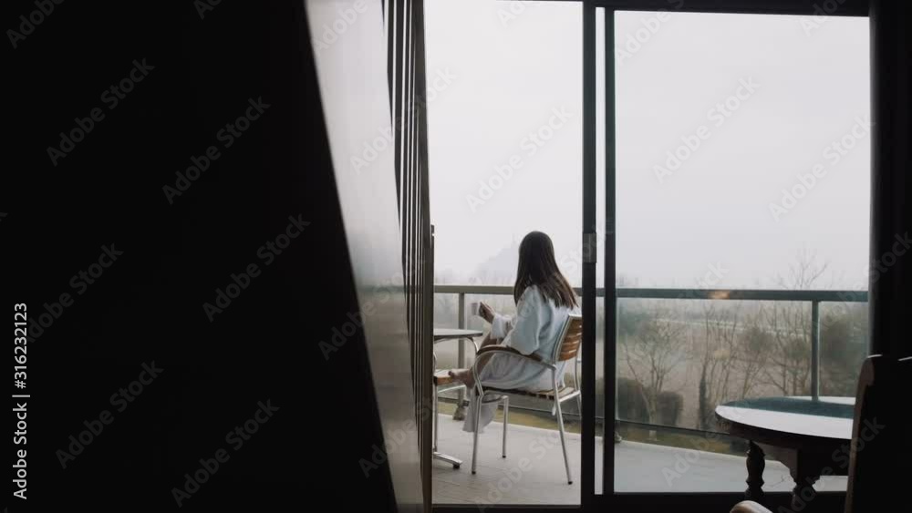 Camera slides right revealing beautiful woman in gown having coffee at room terrace with foggy autumn view slow motion. Camera slides right revealing beautiful woman in gown having coffee at room terrace with foggy autumn view slow motion.