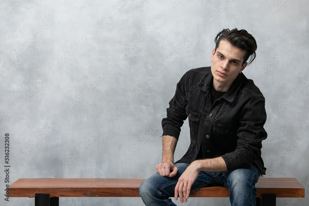 Φωτογραφία Studio fashion portrait of attractive young man in black jacket and blue jeans sitting on wooden bench against grey textured wall