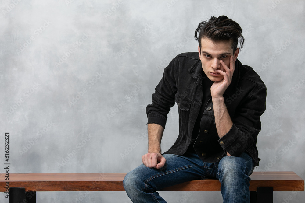 Φωτογραφία Studio fashion portrait of attractive young man in black jacket and blue jeans sitting against grey textured wall