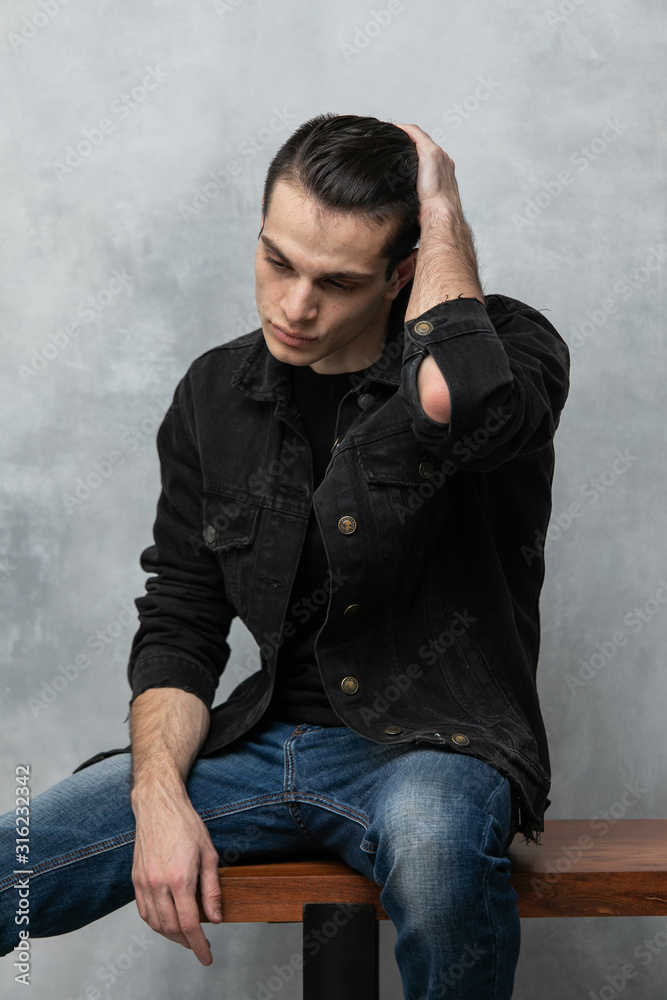Φωτογραφία Studio fashion portrait of attractive young man in black jacket and blue jeans sitting over grey textured wall