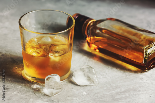 Fotografija Cold whiskey in a glass with ice and a bottle lies on a gray concrete background