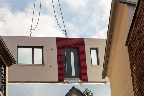 The prefabricated part for a prefabricated house with windows and a red bordered front door is picked up by the truck crane and lifted between two houses for assembly