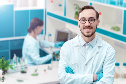 Portrait of smiling man doctor is standing on background of medical laboratory. Scientists are working  making research  conducting experiments  tests with plants. Biologist workplace concept.