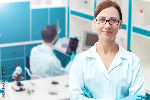 Portrait of smiling woman doctor is standing on background of laboratory. Scientists are working  making research  conducting experiments  tests with plants. Biologist workplace concept.