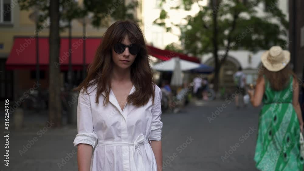 Gimbal shot of woman dressed in white dress and wearing black sunglasses, walking alone on the road on background on Munich pedestrian zone in summer day Gimbal shot of woman dressed in white dress and wearing black sunglasses, walking alone on the road on background on Munich pedestrian zone in summer day