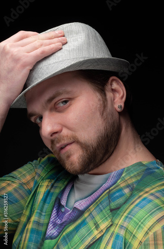 low-key portrait of good-looking bearded serious man with earring in green trendy blazer putting on stylish grey hat isolated on black  © Viktoriia