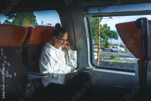 young woman in glasses traveling by train. woman reading newspaper siiting near big window. Enjoying travel time. © Natallia
