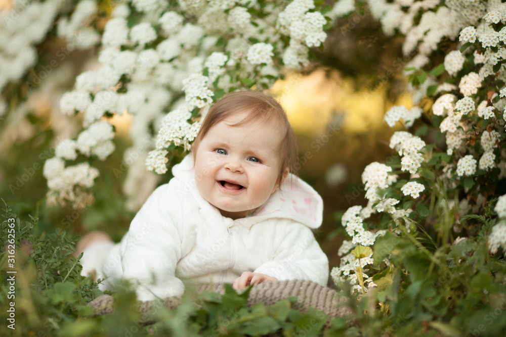 Cute little baby girl in spring in a blooming garden smiling, looking at the camera. Place for the inscription. Concept: spring, family relations, children. The care for the baby. Cute little baby girl in spring in a blooming garden smiling, looking at the camera. Place for the inscription. Concept: spring, family relations, children. The care for the baby.