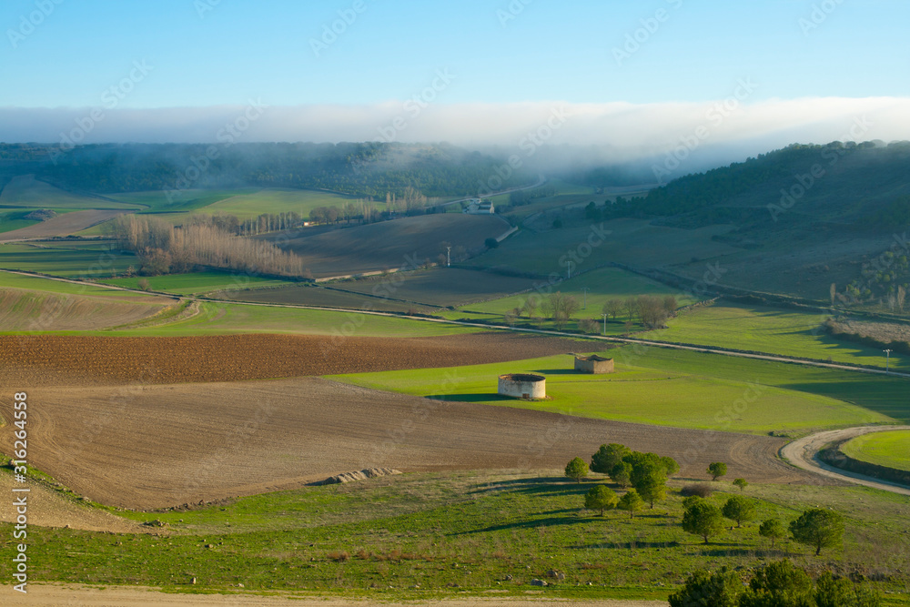 Plowed and green fields in a valley with small buildings, church in the distance and fog in the background