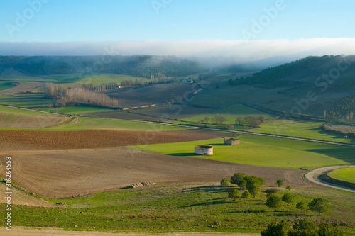 Plowed and green fields in a valley with small buildings, church in the distance and fog in the background