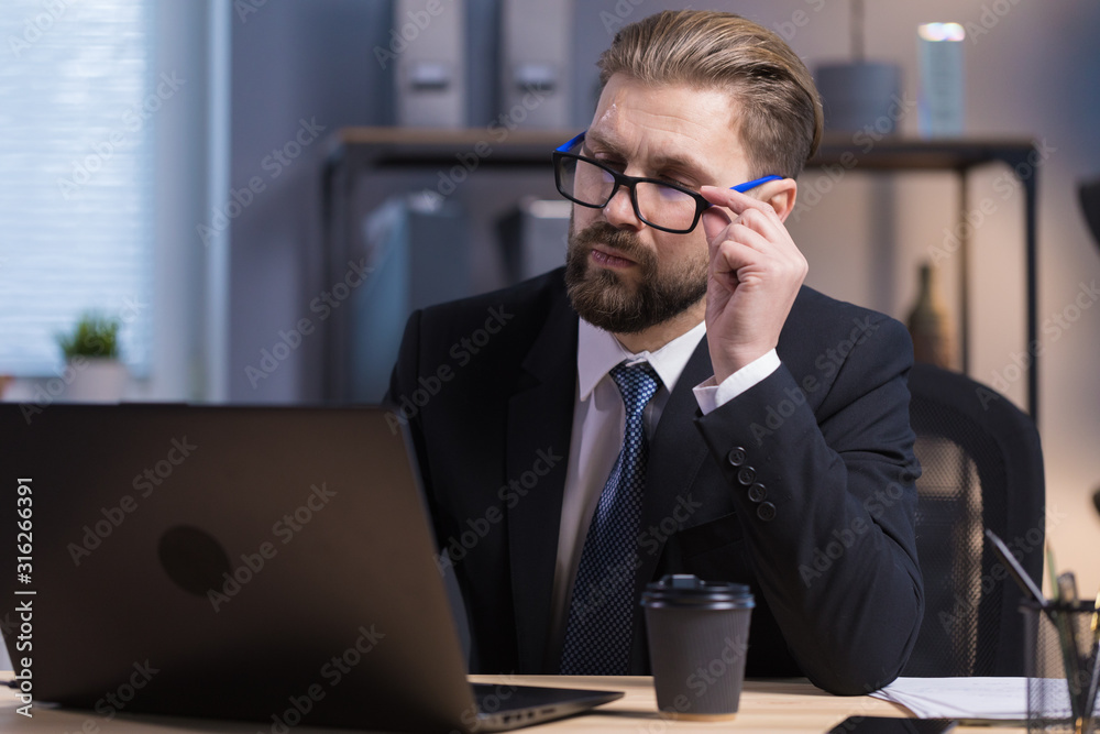 Thoughtful bearded white collar carefully examining data on a laptop screen and adjusting glasses