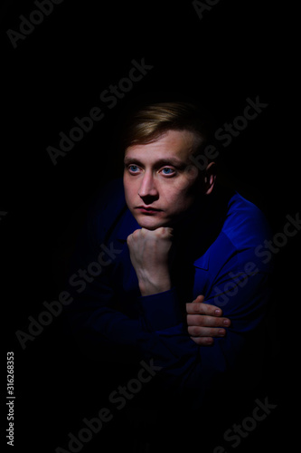 portrait of a handsome young man in a sad shirt sits propping his beard with his hand on a black background
