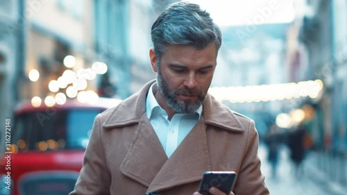 Successful male entrepreneur walking at city central street in downtown and smiling during online messaging via smartphone device slow motion of confident businessman photo