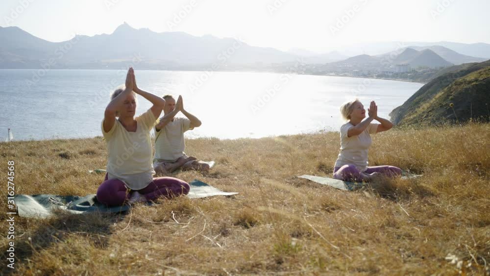Group of senior people doing yoga exercises at the mountain. Elderly man and two woman sitting on the dry grass meadow at sunny day while making asans against sea and mountains on horizon Group of senior people doing yoga exercises at the mountain. Elderly man and two woman sitting on the dry grass meadow at sunny day while making asans against sea and mountains on horizon