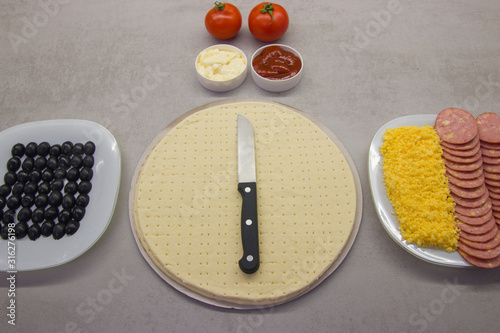pizza preparation process top view: two round blanks of pizza dough with salami olives, cheese on plates with two tomatoes and two sauces next to it on a gray table with texture