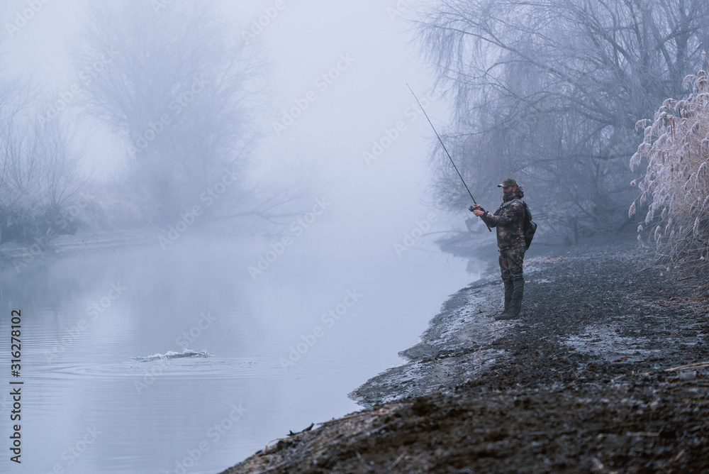 Obraz premium Fisher man fishing with spinning rod on a river bank at misty foggy winter, spin fishing, prey fishing