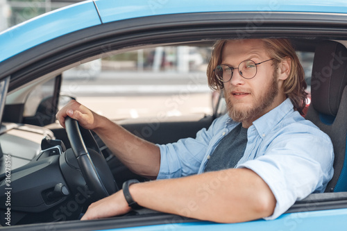 Transportation. Young bearded man in glasses traveling by electric car sitting inside driving looking out the window to camera playful close-up