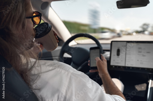 Mode of Transport. Man with long hair in sunglasses traveling sitting inside electric car driving on autopilot drinking coffee browsing smartphone close-up