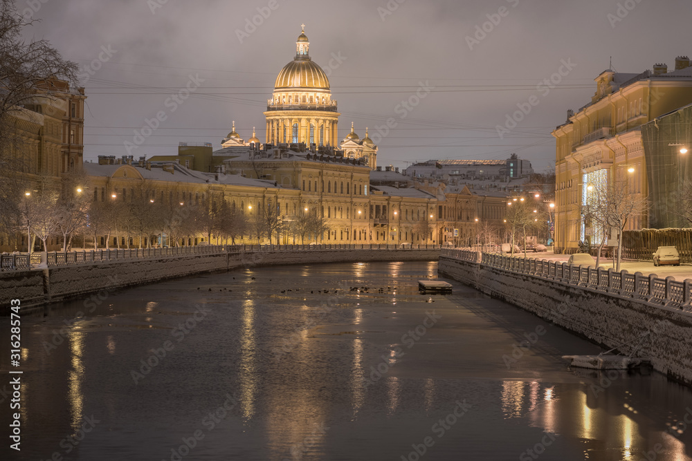 December in Saint Petersburg, Russia. St Isaac Cathedral and the Moyka River after snowfall in the dark early morning. Embankment covered with snow and lit with the dim yellow light of street lamps.