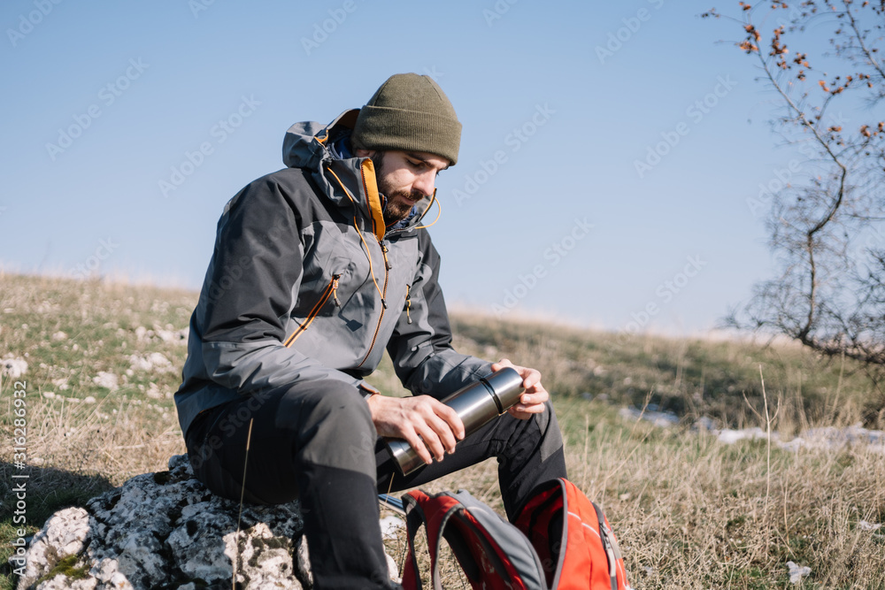 Bearded hiker resting on a stone and holding thermos Bearded hiker resting on a stone and holding thermos