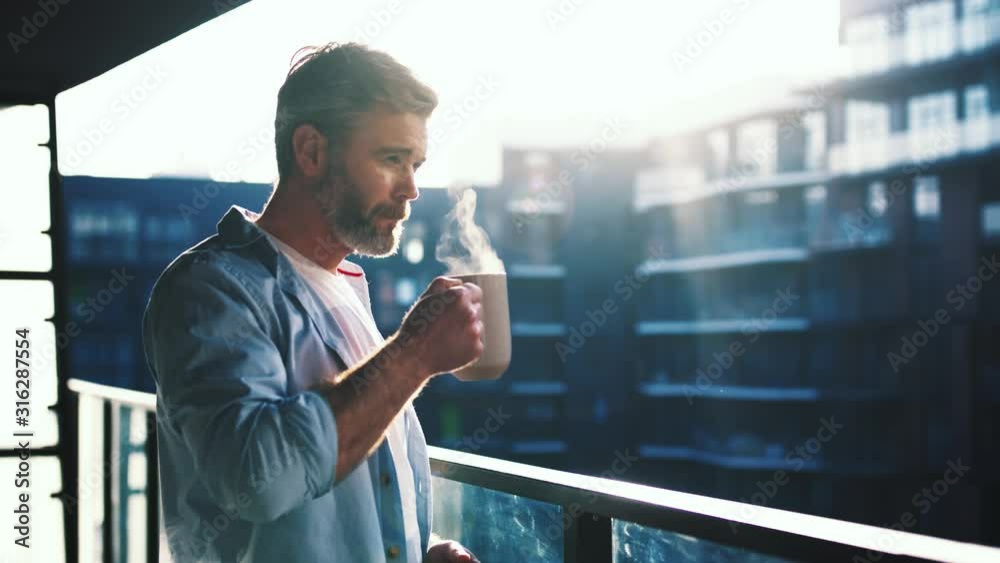 Dreaming european middle aged man stands in the balcony with great views and drinks hot coffee or tea, sun shine, enjoying beautiful morning feeling happy slow motion