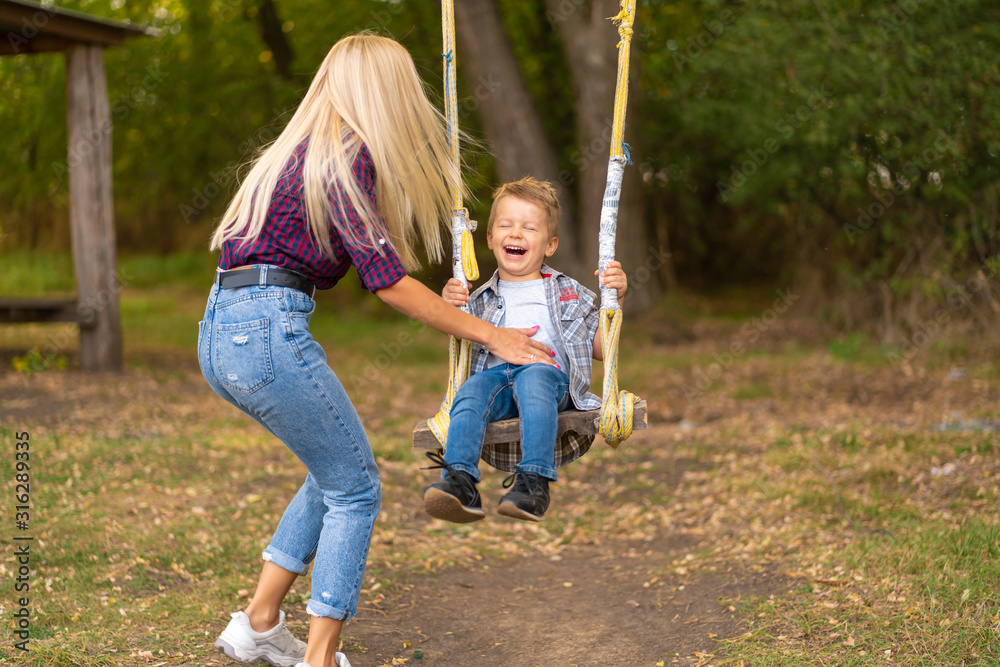 Young blonde mom shakes her little son on a swing in a green park. Happy childhood.