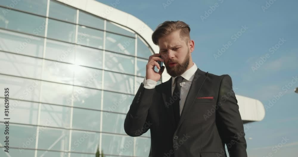 Serious Businessman speaking on his Mobile Phone on morning, Looking at his Wristwatch. Wearing elegant and stylish formal suit. Having well groomed Hairstyle. Huge Glass Building in the Background Serious Businessman speaking on his Mobile Phone on morning, Looking at his Wristwatch. Wearing elegant and stylish formal suit. Having well groomed Hairstyle. Huge Glass Building in the Background