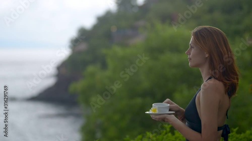 Beautiful red haired woman is drinking tea and watching nature landscape on coast of island. Young happy female enjoying beverage and ocean views while standing on shore with green trees. Concept