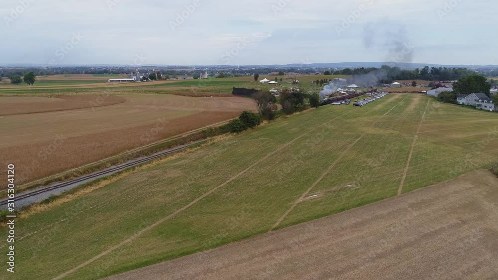 Aerial angled view of a restored steam engine approaching blowing steam and smoke while pulling antique passenger cars with view of farmlands on a sunny day Aerial angled view of a restored steam engine approaching blowing steam and smoke while pulling antique passenger cars with view of farmlands on a sunny day