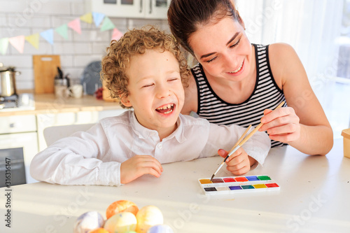 happy young dark-haired mom and her curly blond son paint eggs for easter in the white kitchen in the Scandinavian style