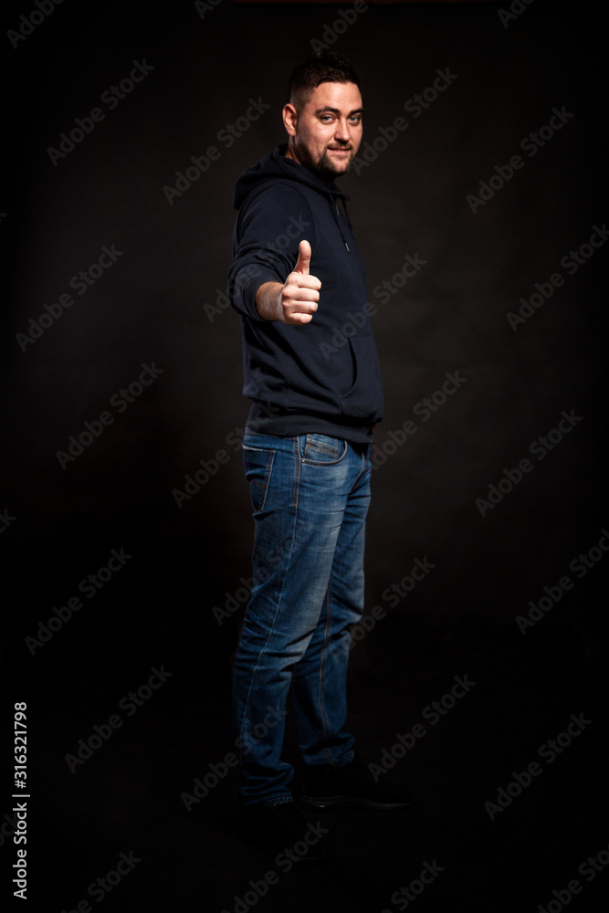 A young man in jeans and a hoodie holds a thumbs up. Full height. Focus in the foreground. Black background. Vertical.