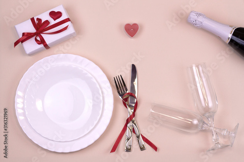 Valentine's Day. Dinner with a table. Plates, cutlery, a gift with a red bow, champagne glasses, a bottle of champagne, hearts on a pink background. View from above.