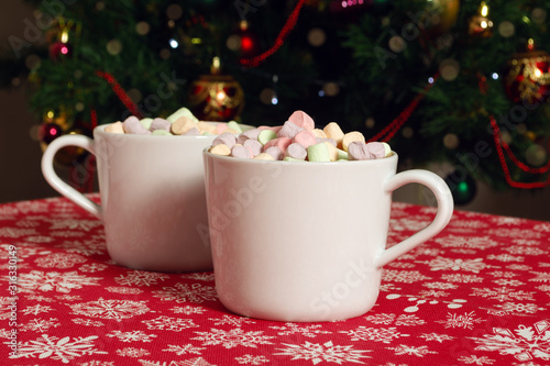 White mugs of hot chocolate beverage with colorful marshmallow on red cloth table with snowflakes on Christmas tree background 