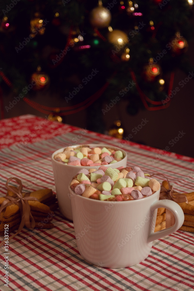 White mugs of hot chocolate beverage with colorful marshmallow and star shaped gingerbread cookies on Christmas tree background White mugs of hot chocolate beverage with colorful marshmallow and star shaped gingerbread cookies on Christmas tree background
