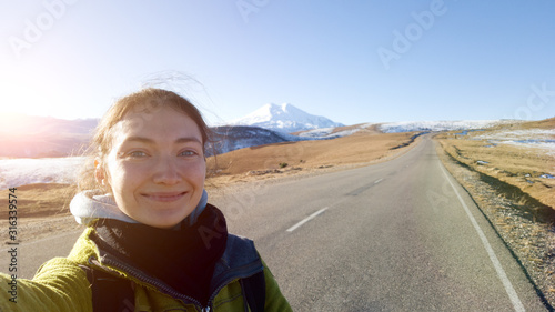 A brunette traveler girl takes a selfie on the background of an asphalt road in the direction of mount Elbrus. Hitchhiking. The glare of the sun. Autumn trip to the Caucasus Russia