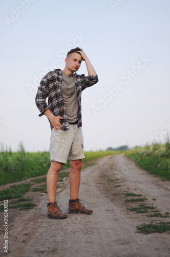 Fototapeta Naklejka Na Ścianę i Meble -  A man in a plaid shirt stands along a dirty road in the countryside.Conceptual photo of traveler.