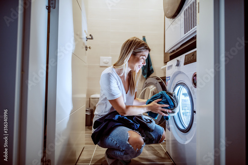 Fotografiet Smiling caucasian blond worthy housewife dressed casual crouching in bathroom and loading washing machine with dirty clothes in late hours