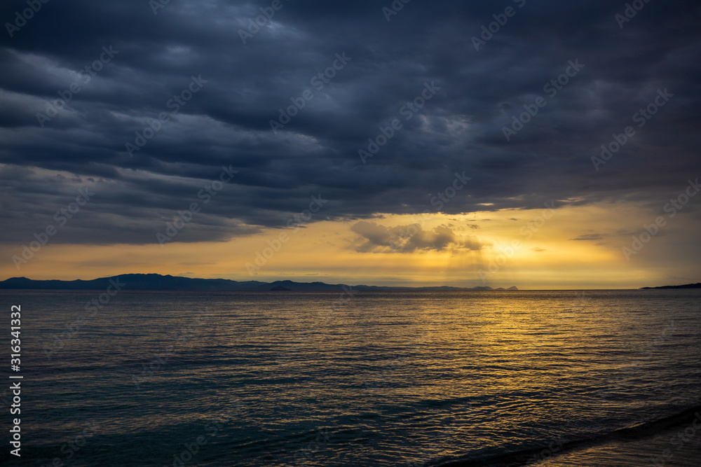 Horizontal color photography of amazing dramatic sunset or sunsrise landscape of dark heavy grey clouds, transparenting through them soft sun beams falling dawn on dark surface of sea water. Greece. 